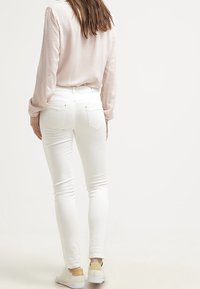 Witte skinny jeans met een medium taille, gecombineerd met een lichtroze blouse met lange mouwen. Gemaakt met witte sneakers met een tan accent.