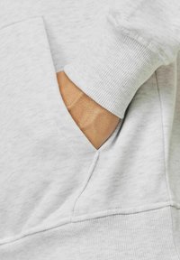Hellgrauer Sweatshirt aus strukturiertem Stoff, mit einer Vorderfronttasche und gerippten Bündchen. Die Tasche zeigt eine Hand, die hinein greift.