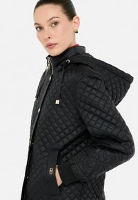 Vrouw in een zwarte gewatteerde jas met capuchon en geborduurde mouwdetails, hand in pocket, kijkend naar de camera.