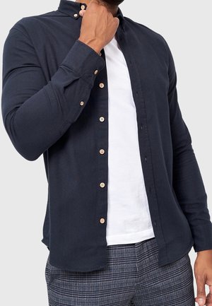 Mies säätämässä tummansinisen, napittamattoman kauluspaidan kaulusta valkoisen t-paidan päällä, harmaat ruutuhousut ja yksinkertainen tausta.