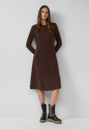 Kobieta z długimi brązowymi włosami nosi ciemnobrązową sukienkę midi z długim rękawem oraz czarne botki na grubych beżowych podeszwach, stoi przy szarej ścianie.