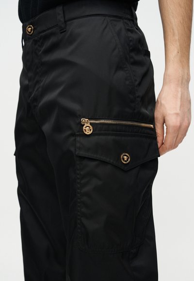 Μαύροι παντελόνια cargo με λεπτομέρειες σε χρυσό φερμουάρ και κουμπί, καπάκι πλαϊνής τσέπης και ορατό χέρι που ακουμπά κοντά στον μηρό.