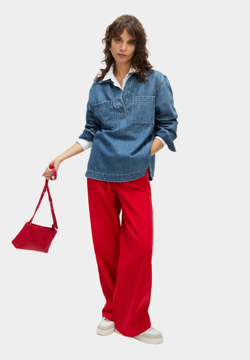 Denim top met lange mouwen en twee borstzakken, gecombineerd met rode wijde broek en een kleine rode handtas. Witte sneakers maken de look af.