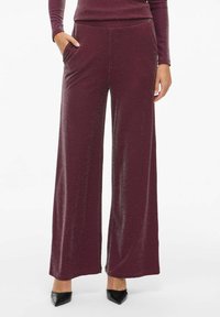 Pantalons bourgogne à jambes larges avec un subtil éclat, portés avec des escarpins noirs à bout pointu, les mains dans les poches de manière décontractée.