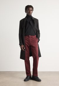 Manteau noir par-dessus un col roulé noir, associé à un pantalon bordeaux et des baskets noires. Tissu lisse avec une coupe ajustée.