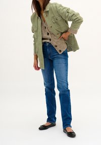 Person wearing blue jeans, black ballet flats, a beige cardigan with large buttons, and an unbuttoned green jacket, standing against a white background.