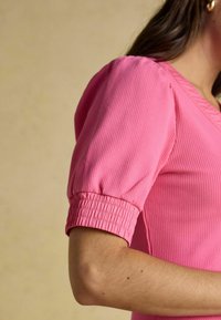 Nahaufnahme einer Person in einem pinkfarbenen, gerippten Oberteil mit gepufften kurzen Ärmeln und elastischen Bündchen vor einem neutralen Hintergrund.