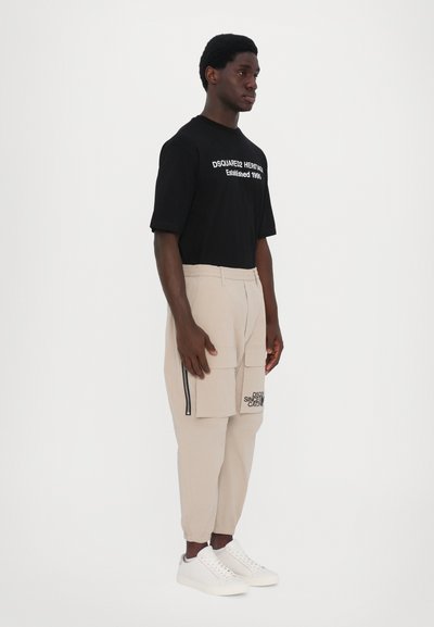 T-shirt noire avec texte blanc, pantalons cargo beiges avec fermetures à glissière et poches, et baskets blanches, présentés sur un modèle debout.