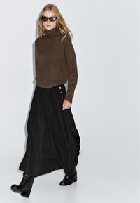 Maglione a collo alto a coste marrone abbinato a una lunga gonna nera con dettagli a bottoni. La modella indossa stivaletti neri alla caviglia e occhiali da sole.