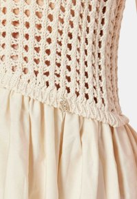 Top de ganchillo en crema con un patrón de tejido abierto, combinado con una falda beige texturizada y fluida. Un acento decorativo es visible en la cintura.