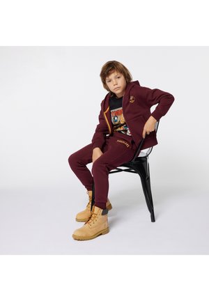 Hoodie bordeaux et joggers avec des détails de logo, associés à des bottes beiges à lacets. Assis sur une chaise noire, avec un fond neutre.