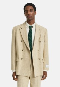 Homme portant un costume beige à double boutonnage avec un pantalon assorti, une chemise blanche et une cravate rayée verte sur fond blanc.