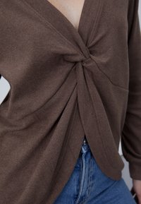 Top marrón de punto con un detalle retorcido en el frente, con escote en V y mangas largas, combinado con jeans de mezclilla azul.