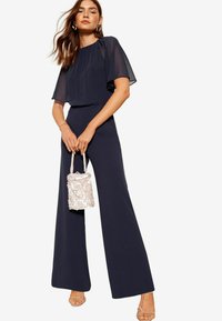 Mono azul marino con perneras anchas y un top corto de manga corta transparente, acompañado de un pequeño bolso texturizado en rosa claro.