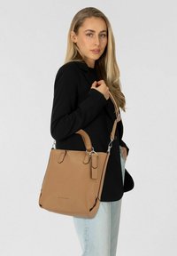 Tan leren handtas met textuur, twee handvatten en een afneembare schouderriem. Model draagt een zwarte blazer en lichte jeans.