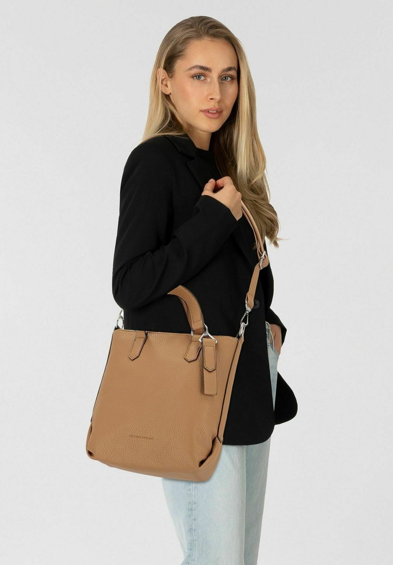 Tan leren handtas met textuur, twee handvatten en een afneembare schouderriem. Model draagt een zwarte blazer en lichte jeans.