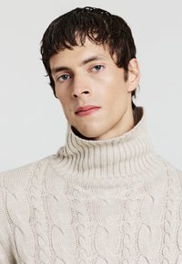 Beige gestrickter Rollkragenpullover mit strukturierten Zopfmustern und einem gerippten Kragen. Weicher, dehnbarer Stoff mit einer taillierten Passform.