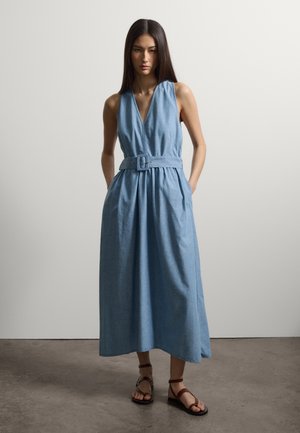 Frau mit langen dunklen Haaren, die ein ärmelloses blaues Midikleid mit Gürtel und braune flache Sandalen mit Riemen trägt, steht auf einem Betonboden.