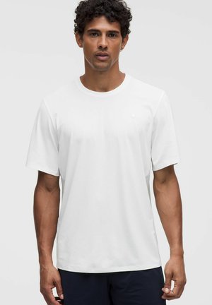 Mann mit lockigem Haar, der ein schlichtes weißes T-Shirt mit kurzen Ärmeln trägt, steht vor einem hellgrauen Hintergrund.