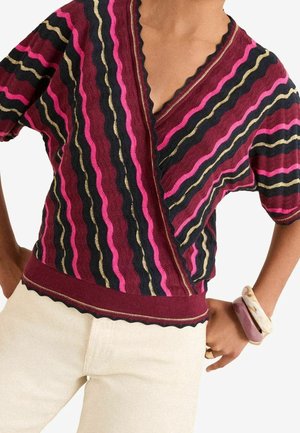 Kvinde iført en kortærmet top i bordeaux, pink, sort og guld med stribede teksturer og bølgekant, beige bukser og chunky armbånd.