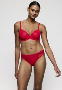 Vrouw met een gemiddelde huidtint, gekleed in een rode beha en bijpassend ondergoed, staand tegen een effen lichte achtergrond, zacht glimlachend.