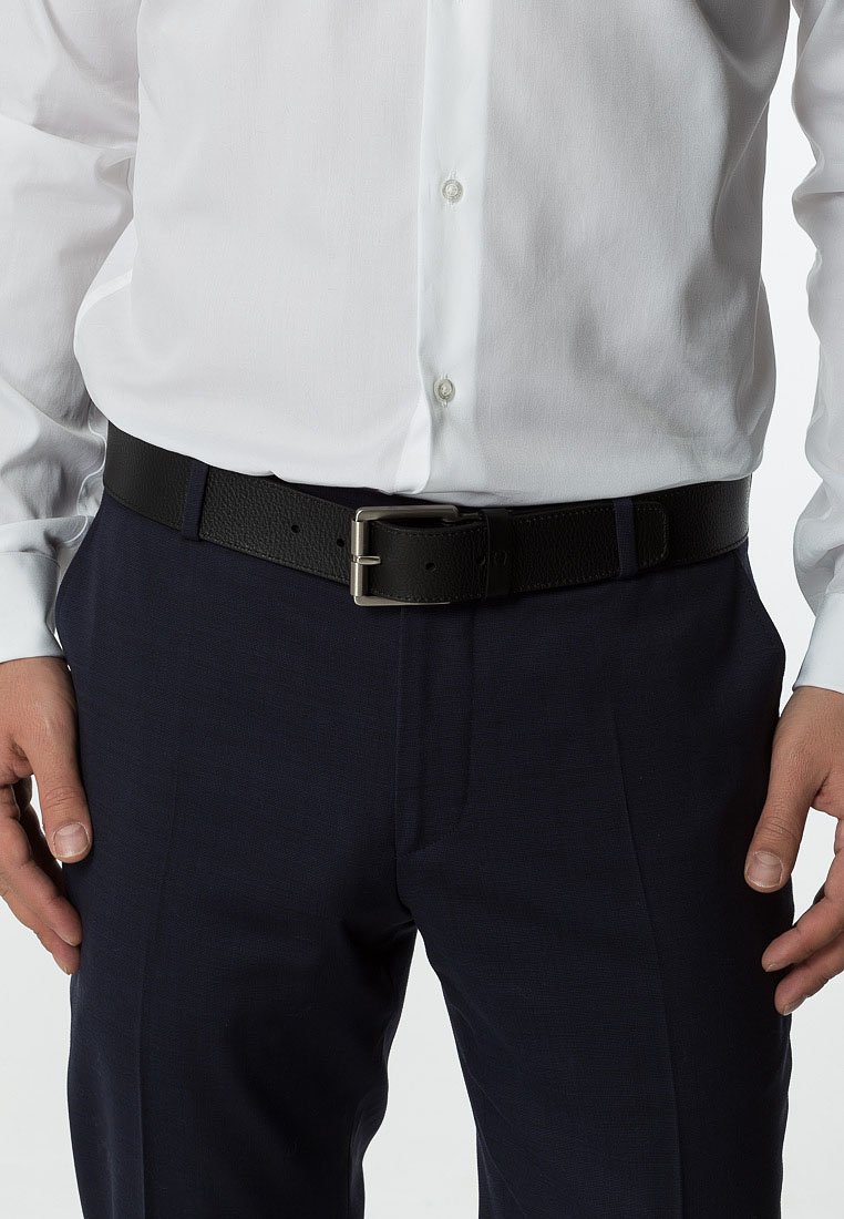 Cinturón de cuero negro con un acabado texturizado y una hebilla de metal plateado. Lleva puesto con una camisa blanca y pantalones oscuros.