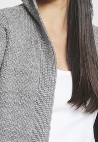 Szary dziergany kardigan z kapturem, o fakturowanym wzorze. Tkanina wydaje się miękka, noszony na białej koszuli, z widocznymi długimi ciemnymi włosami.