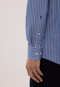 Niebiesko-biała koszula w paski z białymi guzikami i detalem na mankiecie, wykonana z gładkiej tkaniny, ukazująca część dłoni.