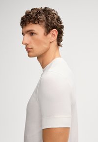 Junger Mann mit lockigem braunem Haar, der ein eng anliegendes weißes Kurzarmhemd trägt und nach links blickt, vor einem schlichten hellen Hintergrund.