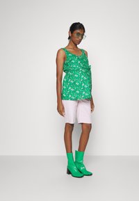 Top verde floral com um detalhe em nó, combinado com calções rosa claro e botas de tornozelo verdes brilhantes. Modelo a usar óculos de sol verdes oversized.