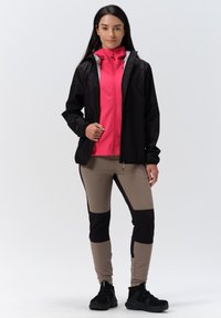Svart regnjacka med hög krage över en rosa zip-up topp. Beige och svarta panelbyxor, kombinerat med svarta träningsskor.