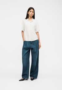 Γυναίκα που στέκεται με τα χέρια στις τσέπες φορώντας λευκό κοντομάνικο πουκάμισο, χαλαρά, σκούρα μπλε φαρδιά τζιν και μαύρα μυτερά παπούτσια.