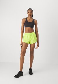 Sujetador deportivo negro, pantalones cortos neon verdes, cinturilla elástica, detalle de logo reflectante. Modelo vistiendo zapatillas deportivas negras, de pie contra un fondo blanco.