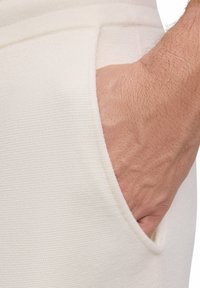 Roomkleurig textiel met een zichtbare zijzak, waar een hand in rust; het materiaal lijkt zacht en geweven.