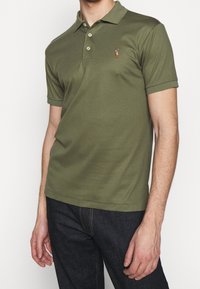 Olivgrön polo med kort ärm, tillverkad av bomull, med en tre-knapps knäppning och en liten broderad logotyp på vänster bröst.