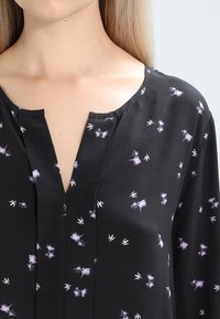 Černá blůza s výstřihem do v a měkkou tkaninou, pokrytá květinovým vzorem v purpurové a bílé barvě. Krátké rukávy, lehká textura.