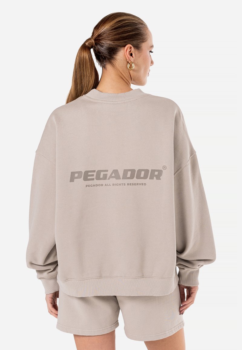 Beige oversized hettegenser med fallende skuldre, ribbede mansjetter og "PEGADOR"-logo trykket på baksiden. Laget av myk stoff.