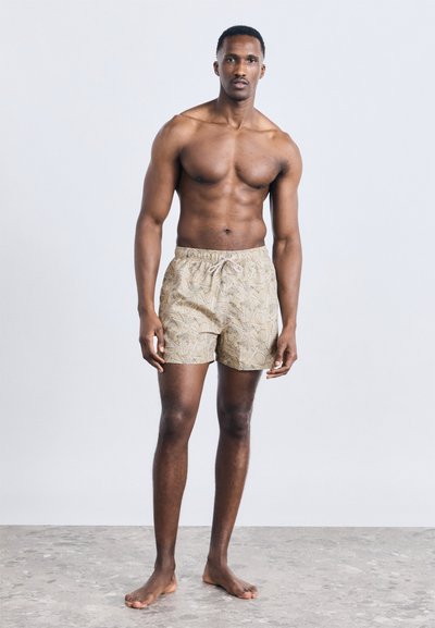 Άνδρας χωρίς πουκάμισο με μυώδες σώμα, όρθιος ξυπόλητος σε πέτρινο δάπεδο, φοράει μπεζ μαγιό με σχέδιο και κορδόνι, μπροστά σε απλό φόντο.