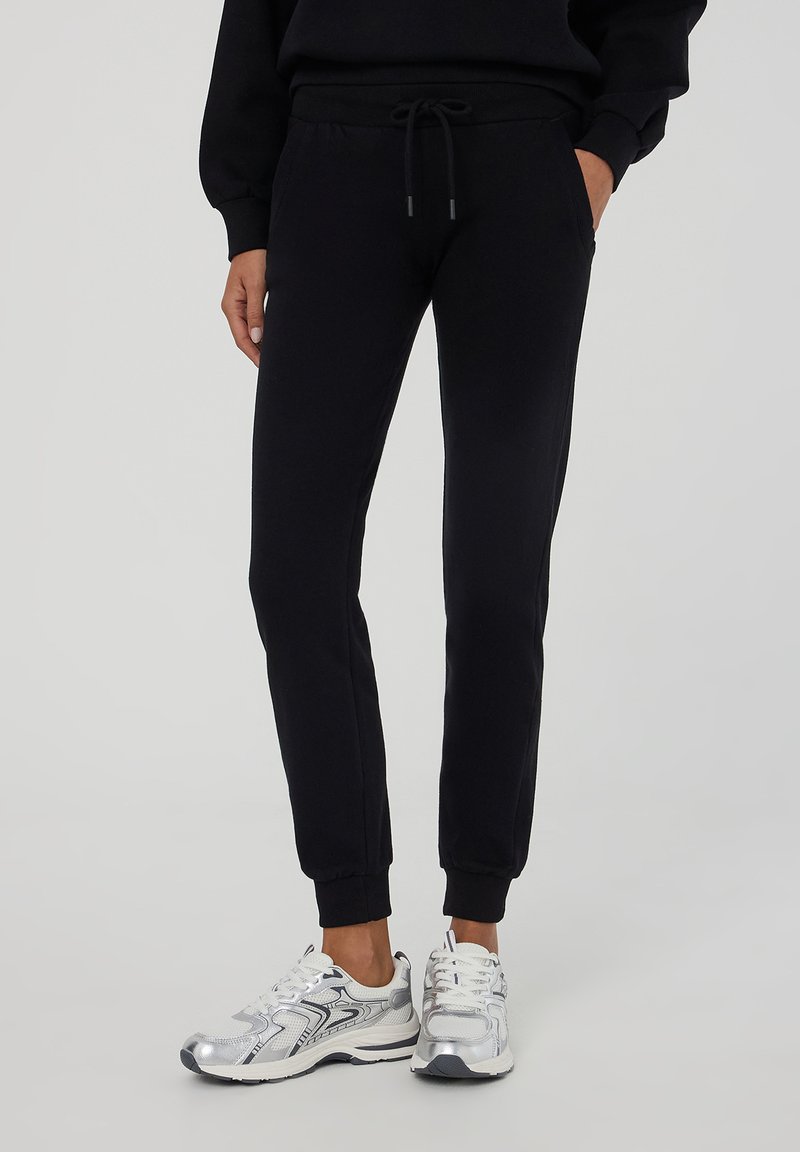 Pantaloni sportivi neri con polsini elastici, vita con cordoncino e tasche laterali, realizzati in tessuto morbido, abbinati a sneakers sportive argento.