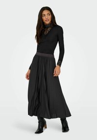 Gonna maxi plissettata nera con una vita scura, abbinata a un top aderente a maniche lunghe con un colletto in pizzo strutturato e polsini.