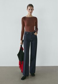 Brązowa dopasowana bluzka z długim rękawem zestawiona z ciemnymi dżinsami o wysokim stanie i szerokich nogawkach, uzupełniona czarno-czerwoną torbą oraz czarnymi butami.