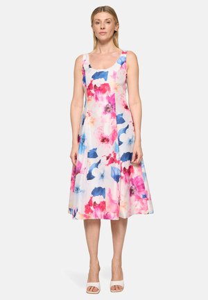 Kvinde iført hvid ærmeløs kjole med pink, rød og blå blomstermønster, stående med afslappede arme, iført hvide åbne hæle.