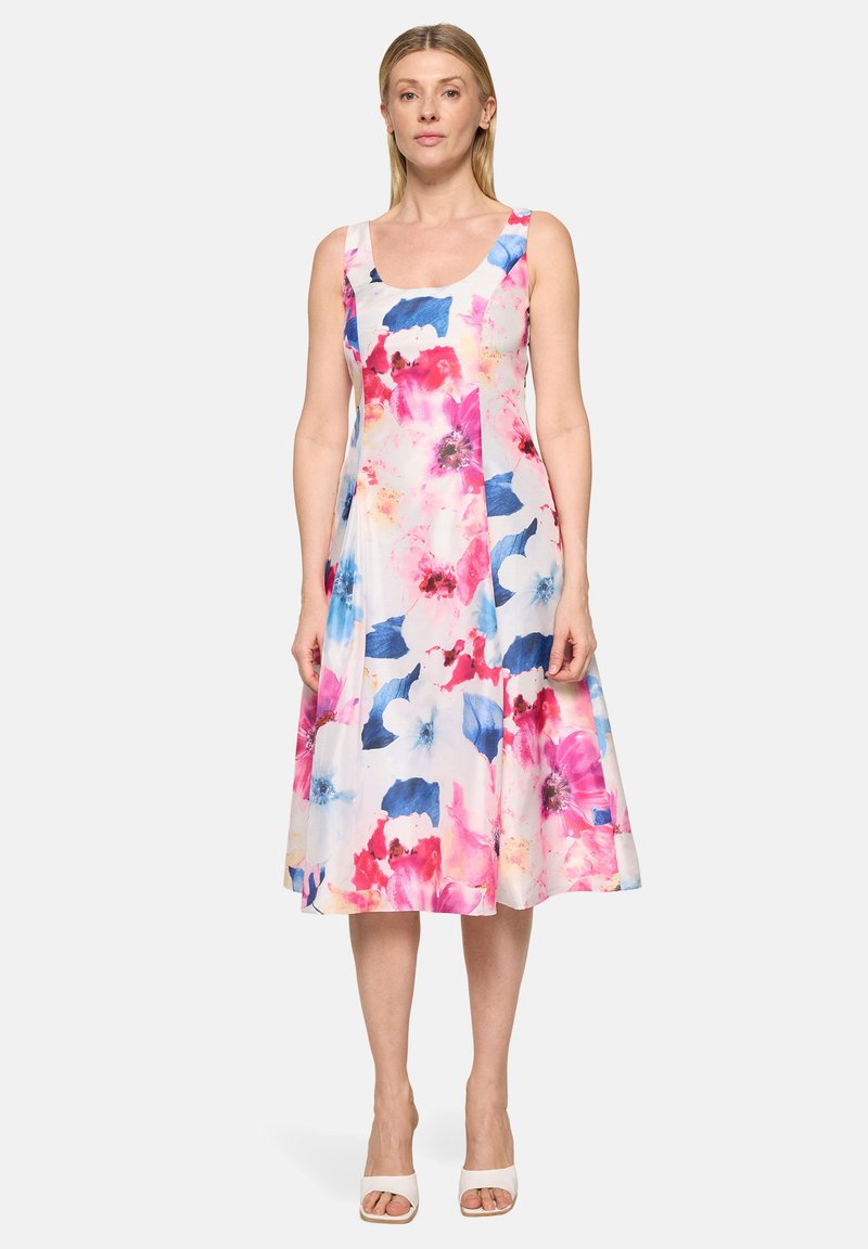 Femme portant une robe blanche sans manches avec un imprimé floral rose, rouge et bleu, debout les bras détendus, portant des talons ouverts blancs.