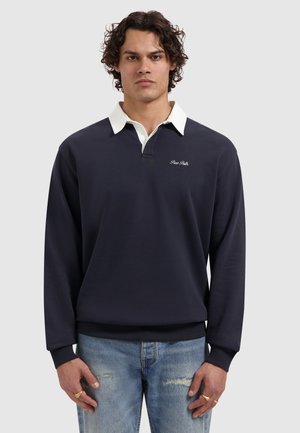 Man met krullend haar, gekleed in een marineblauwe sweatshirt met een witte kraag, lichtblauwe gescheurde spijkerbroek, en gouden ringen aan beide handen.