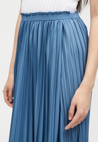 Γυναίκα που φοράει μια μπλε πλισέ φούστα με ψηλή μέση και βολάν στη μέση, μαζί με λευκό πλεκτό τοπ, σε απλό φόντο.