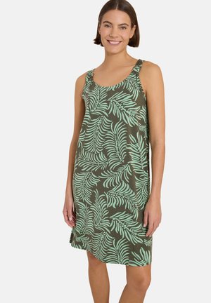 Femme souriante portant une robe verte sans manches avec un motif de feuilles tropicales vert clair, debout devant un fond clair uni.