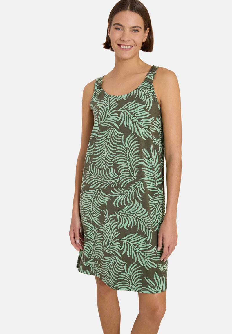 Femme souriante portant une robe verte sans manches avec un motif de feuilles tropicales vert clair, debout devant un fond clair uni.