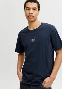 Camiseta de algodón azul marino con mangas cortas y cuello redondo, que presenta un logo blanco y púrpura en el pecho. Textura suave.