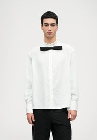 Bílá košile s dlouhým rukávem a volánkovým límcem, doplněná černým motýlkem. Černé kalhoty, hladká tkanina a uvolněný střih.