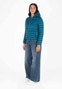 Giacca puffer color teal con cappuccio, caratterizzata da un design a cuciture orizzontali e una zip dorata. Abbinata a jeans larghi di colore blu e sneakers bianche.
